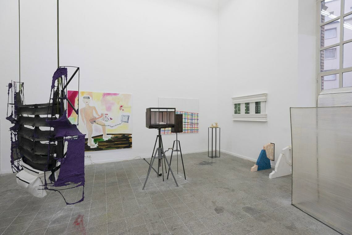 Foto: Ein Raum mit weißen Wänden, grauem gefließten Boden und einem Fenster. An den Wänden hängen mehre flache Objekte im Groß- und Mittelformat. Im Raum stehen Skulpturen. Von der Decke hängt ein weiteres Objekt.