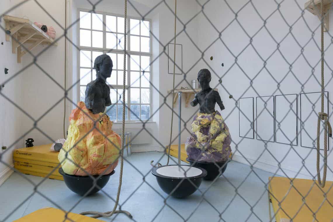 Foto: Ein Raum mit weißen Wänden, grauem Boden und einem großen Fenster. An der Wand des Raumes befinden sich, auf unterschiedlichen höhen, flache, gerahmte Objekte und zwei regalbrettartige Konstruktionen, auf denen Objekte liegen. Auf dem Boden liegen Stapel gelber Matten. Im Raum stehen auérdem drei große Skulpturen. Von der Decke hängen Seile. Durch den Raum ist ein Gitter gespannt, durch welches das Foto aufgenommen wurde.