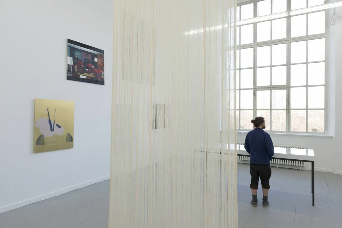 Foto: Ein Raum mit weißen Wänden, grauem Boden und einem großen Fenster. An der Wand hängen auf unterschiedlicher Höhe mehre flache Objekte. Von der Decke hängt ein gelblicher Vorhang auf beigen, schmalen Fäden. Im Raum steht ein Tisch mit einer weißen Tischplatte. Vor dem Tisch steht ein Mensch.