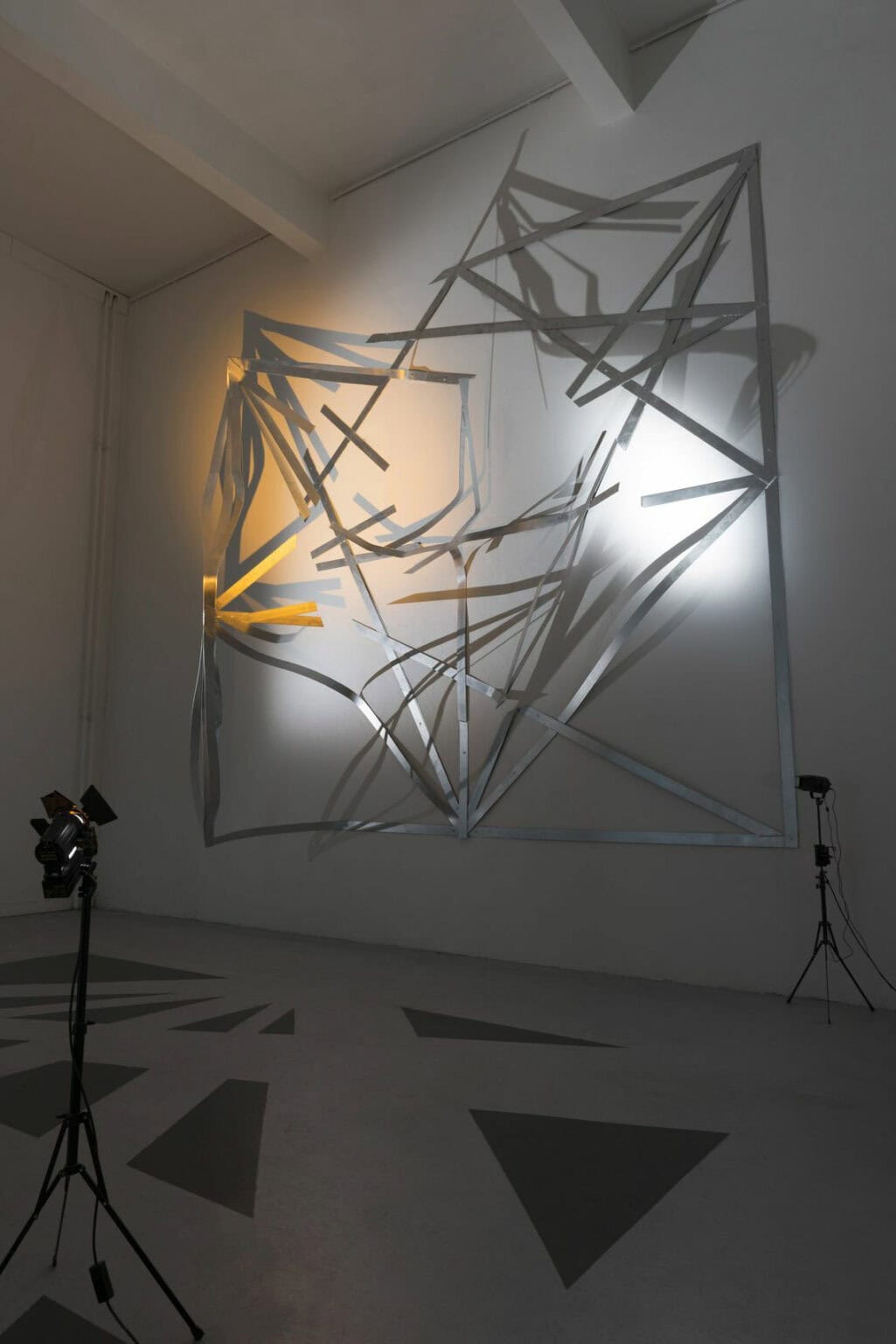 Foto: Ein Raum mit weißen Wänden und grauem Boden. Im Raum stehen zwei an Stativen montierte Lichter, die auf eine große, flache Metallkonstruktion gerichtet sind, die an der Wand hängt. Auf dm Boden vor der Konstruktion sind in dunkelgrau dunkelgraue Dreiecke aufgemalt.