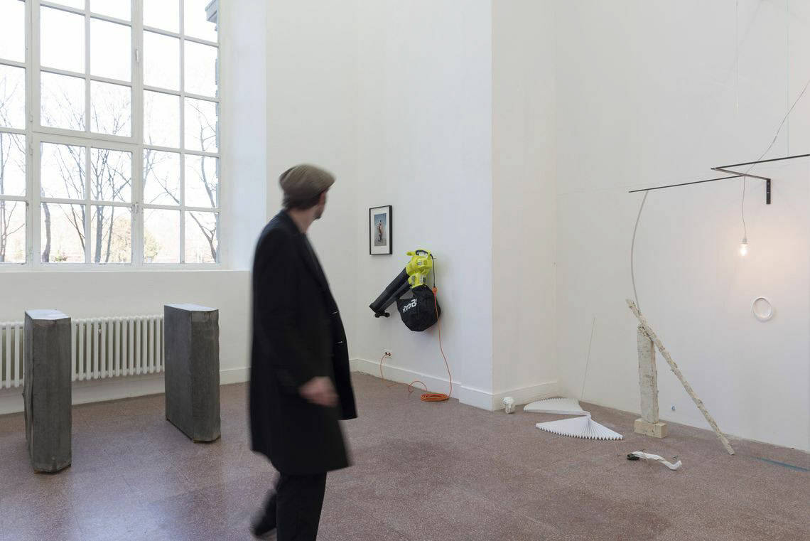 Foto: Ein Raum mit weißen Wänden, rotem Steinboden, einem großen Fenster und einer wandhohen, bogenförmigen Einbuchtung. An den Wänden und auf dem Boden des Raumes befindet sich eine Vielzahl von Objekten. Durch den Raum bewegt sich ein Mensch.