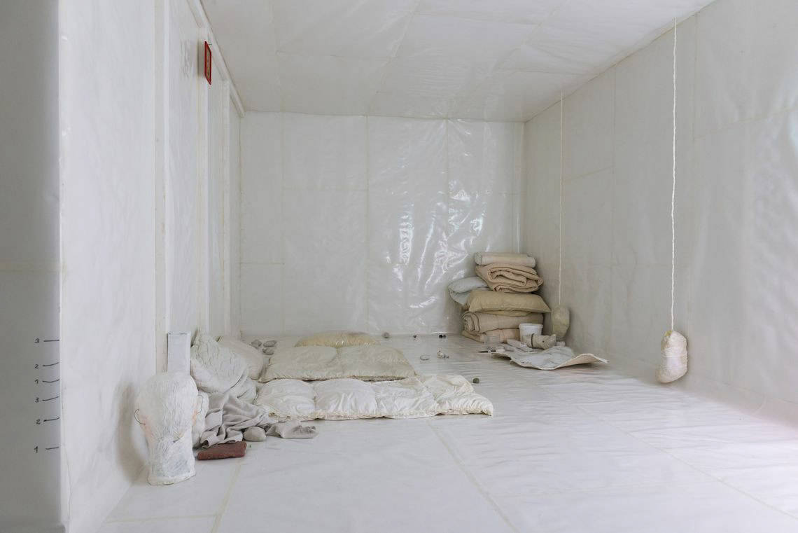Foto: Ein Raum, der komplett mit einem Material in weißer Farbe ausgekleidet ist, dass eine plastikartige, reflektierende Oberfläche hat. Im Raum befinden sich viele Objekte, darunter gefaltete und ausgebreitete Textilien, die zu einer Art Bettenlager angeordnet sind. Von der Decke hängen weitere Objekte bis kurz über den Boden.