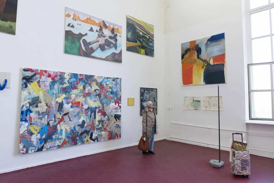 Foto: Foto: Ein Raum mit weißen Wänden, dunkelrotem Boden und einem großen Fenster. An der Wand befinden sich mehrere flache Objekte im Klein-, Mittel- und Großformat, die unter- und nebeneinander auf unterschiedlicher Höhe hängen. Im Raum stehen Skulpturen auf dem Boden. Im Raum befindet sich ein Mensch.