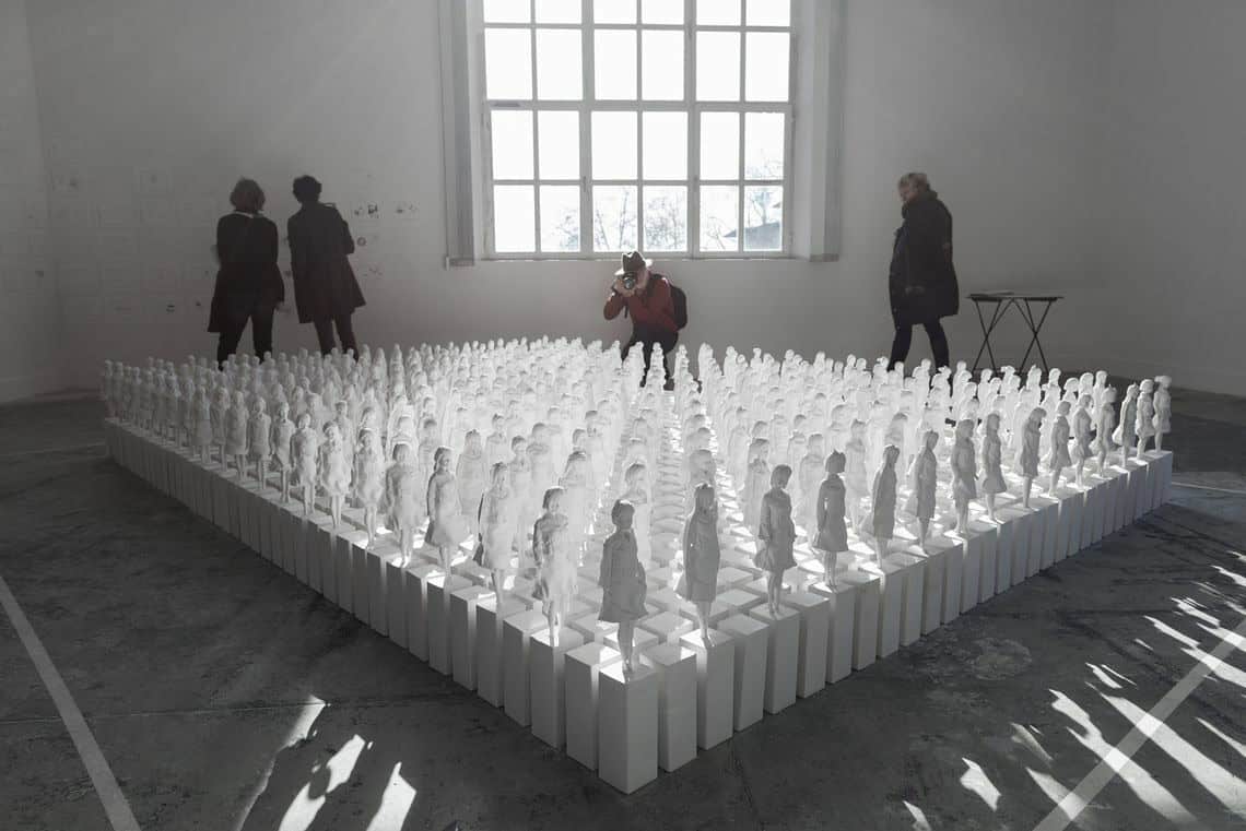 Foto: Ein Raum mit weißen Wänden, grauem Boden und einem großen Fenster. Im Raum steht eine große Skulptur, die auf einer Gruppe nah beieinander stehender Objekte besteht. Sonnenlicht fällt auf das Objekt, so dass dieses einen Schatten wirft. An der Wand hängen mehrere Papiere. Durch den Raum bewegen sich Menschen.