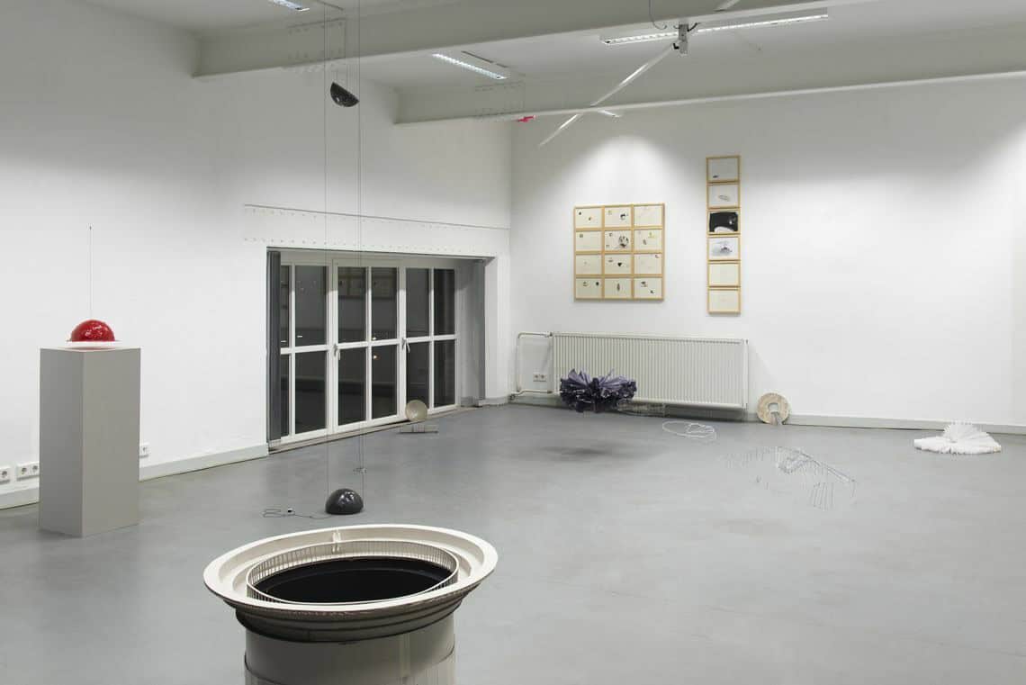 Foto: Ein räum mit weißen Wänden, grauem Boden und einem kleinen Fenster. An der Wand hängen zwei Gruppen gerahmter Objekte, die einen in einer Linie untereinander und die anderen zu einem Quadrat angeordnet. Im Raum verteilt stehen und liegen Objekte auf dem Boden. Von der Decke hängen weitere Objekte von Schnüren.