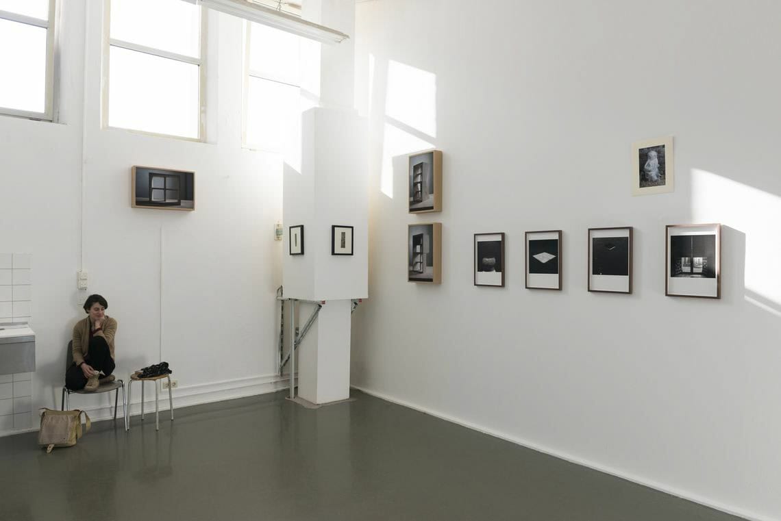 Foto: Ein Raum mit weißen Wänden und grauem Boden. Oben um Raum eine Reihe aus Fenstern. An der Wand hängen, auf unterschiedlicher Höhe, mehrere gerahmte Objekte in einem ähnlichen Format. In dem Raum befindet sich eine säulenartige Verkofferung, auf der ebenfalls kleine, gerahmte Objekte hängen. Links im Raum stehen zwei Hocker. Auf einem der Hocker sitzt eine Person.