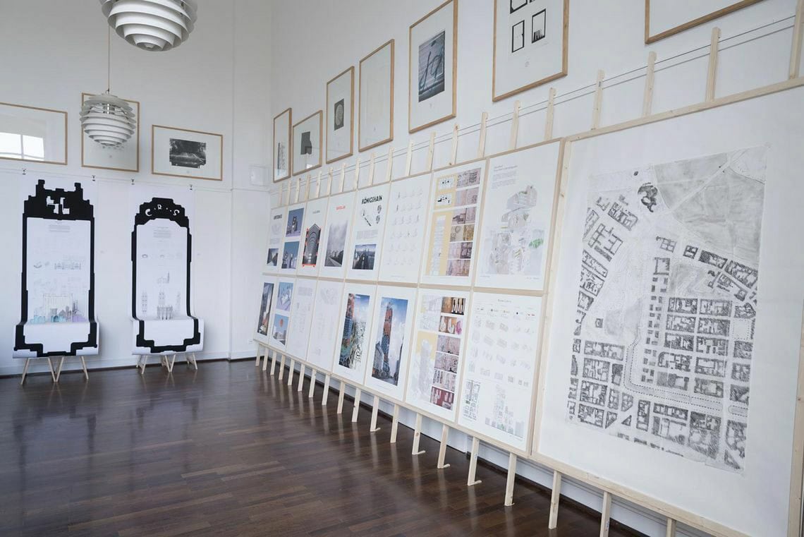 Foto: Ein Raum mit weißen Wänden und braunem, poliertem Dielenboden. Oben an der Wand hängen, in einer Reihe auf gleicher Unterkante, gerahmte Objekte. Darunter befindet sich eine Konstruktion aus Holzleisten, die auf dem Boden aufliegt und an die Wand lehnt. An die Leisten sind gerahmte Objekte montiert. An einer anderen Wand hängen Papierrollen, die auf sich nah an der Wand befindliche Objekte herunterreichen, die durch das herabfallende Papier verdeckt werden.