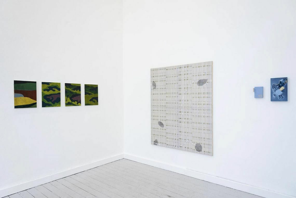 Foto: Die Ecke eines Raume mit weißen Wänden und grauem Dielenboden. An den Wänden hängen flache Objekte in unterschiedlichen Formaten.