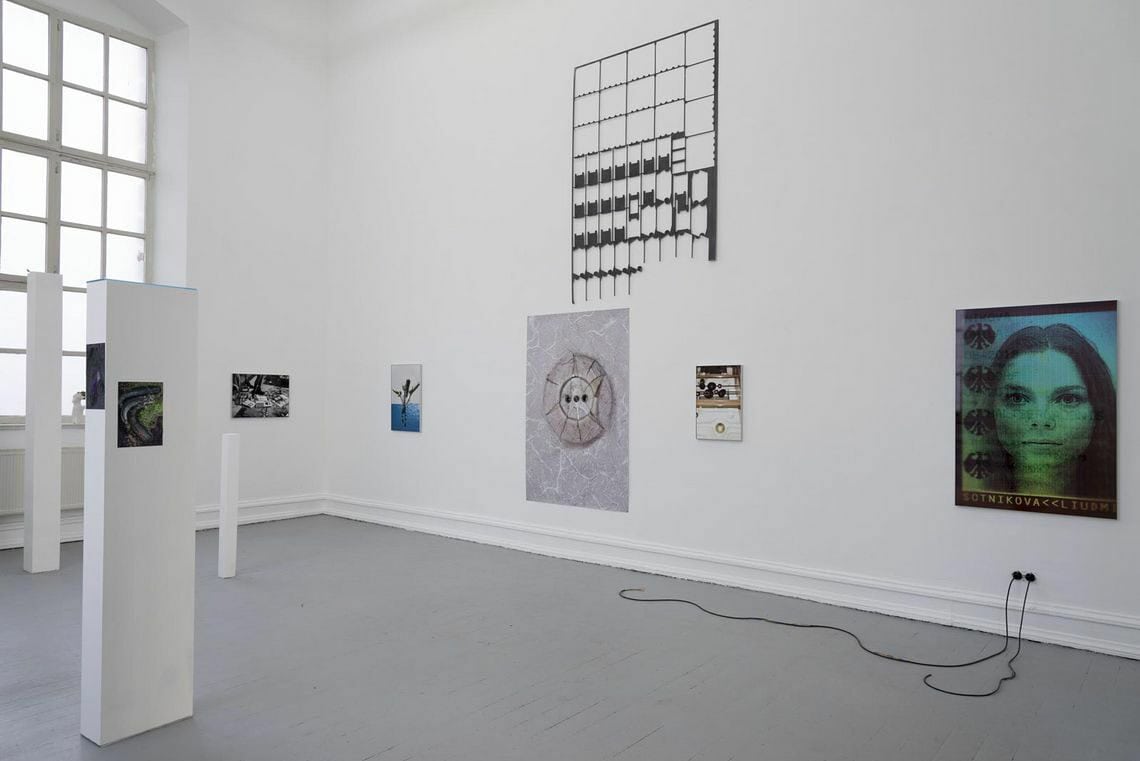 Foto: Ein Raum mit weißen Wänden, grauem Dielenboden und einem Fenster. An der Wand befinden sich auf unterschiedlicher Höhe mehrere Objekte aus unterschiedlichen Materialien. Im Raum befinden sich mehrere schmale Säulen in unterschiedlichen Höhen und Formaten. An den Säulen hängen ebenfalls flache Objekte.