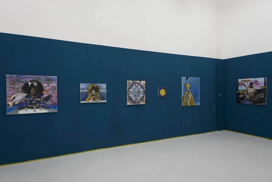 Foto: Ein Raum mit einem brauten Streifen Blauer Farbe an der Wand. Die darüber befindliche Wand ist Weiß. Der Boden ist, bist auf einen kleinen gelben Streifen in Wandnähe, grau. An den Wänden hängen, auf gleicher Mittellinie, klein- und mittelformatige Leinwandarbeiten.