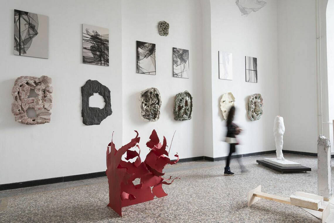 Foto: Ein Raum mit weißen Wänden und mehrfarbigem Steinboden. An der Wand befinden sich mehrere Objekte, die in zwei Reihenim gleichen Abstand zueinander hängen. Im Raum verteilt befinden sich mehrere Skulpturen aus unterschiedlichen Materialien. Durch den Raum bewegt sich ein Mensch.