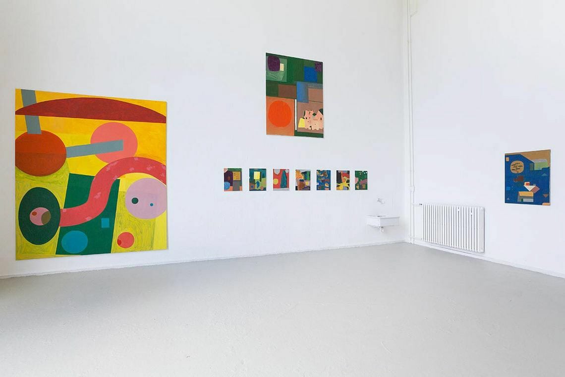 Foto: Ein Raum mit weißen Wänden und grauem Boden. An den Wänden hängen Leinwandarbeiten im Klein-, Mittel- und Großformat auf unterschiedlicher Höhe.