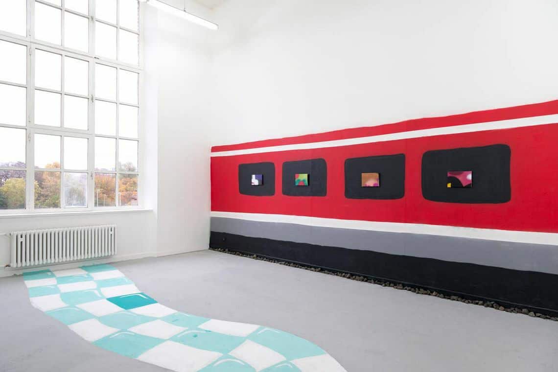 Foto: Ein Raum mit weißen Wänden, grauem Boden und einem großen Fenster. Teile der Wand und die Fußleisten sind mit unterschiedlichen breiten Bahnen aus schwarzer, grauer und roter Farbe bedeckt. Auf dem streifen roter Farbe sind Schwarze Rechtecke aufgemalt. Innerhalb der schwarzen Rechtecke hängen flache, rechteckige Objekte. auch Teile des Bodens sind mit einem gewelltem Pfad bemalt, der ein blau-weißes Schachbrettmuster hat.