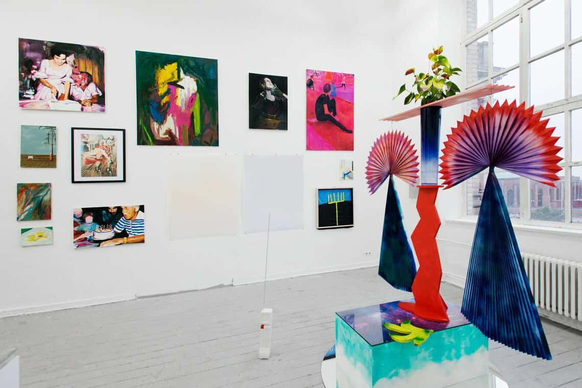 Foto: Ein Raum mit weißen Wänden, grauem Dielenboden und einem großen Fenster. An den Wänden hängen mehrere flache Objekte neben- und untereinander. Manche von Ihnen sind gerahmt. Im Raum Stegen Skulpturen, die aus unterschiedlichen Materialien bestehen.