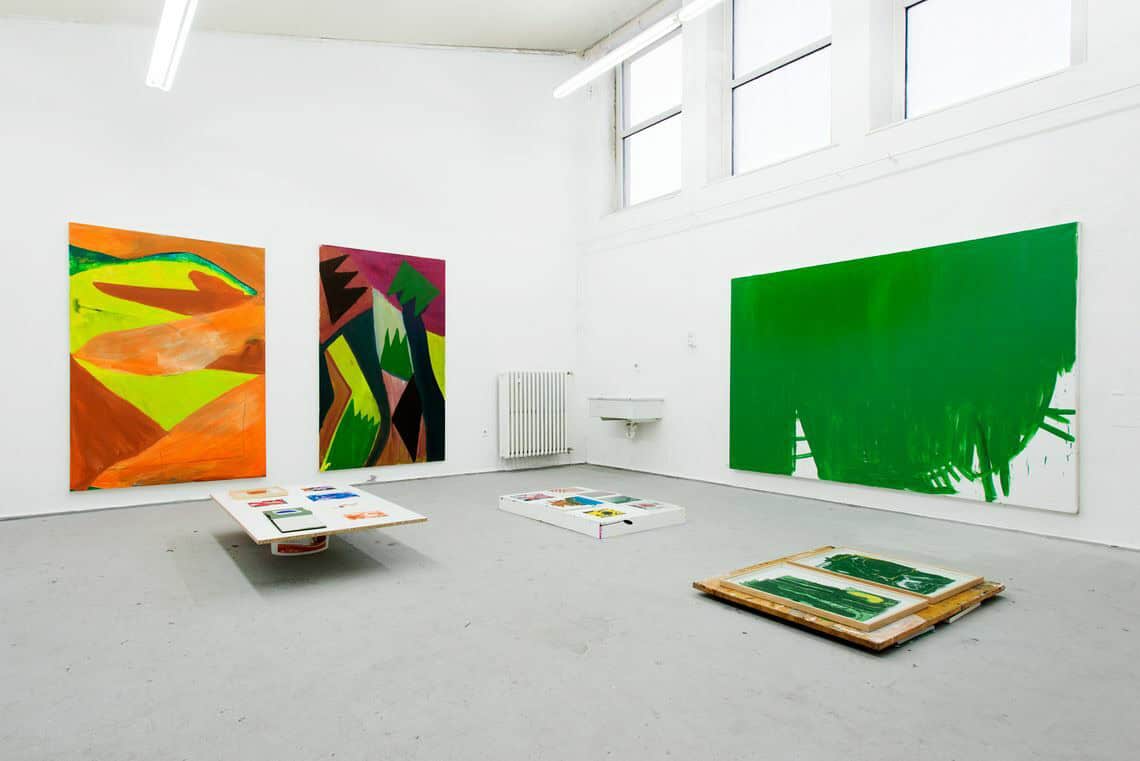 Foto: Ein Raum mit weißen Wänden und grauem Boden. Oben An der Wand eine leiste auf Fenstern. An der Wand hängen großformatige Leinwandarbeiten. Auf dem Boden liegen flache Objekte.