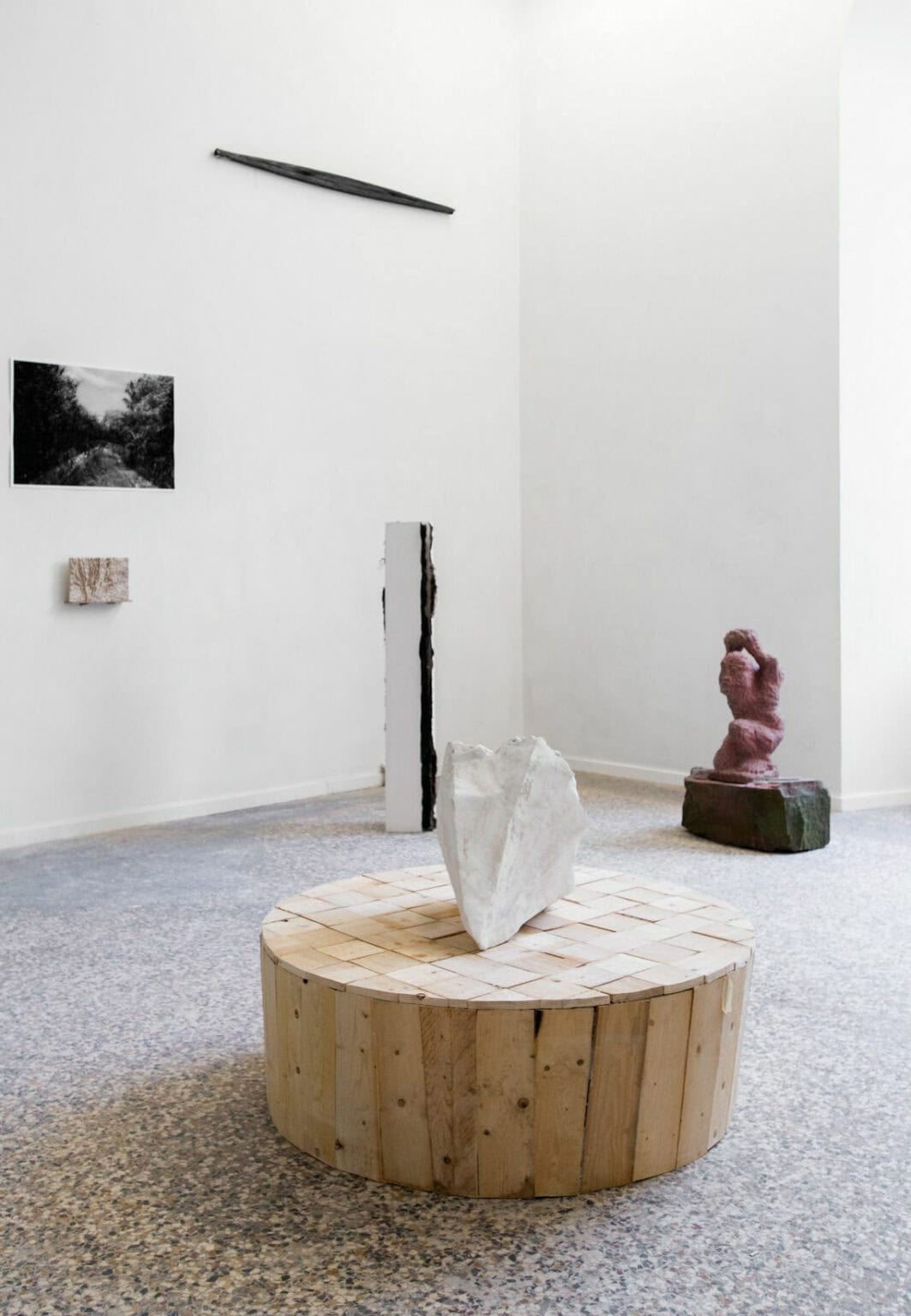 Foto: Ein Raum mit weißen Wänden und mehrfarbigem Steinboden. An den Wänden befinden sich flache Objekte, die auf unterschiedlicher Höhe hängen. Im Raum befinden sich mehrere Skulpturen aus unterschiedlichen Materialien.