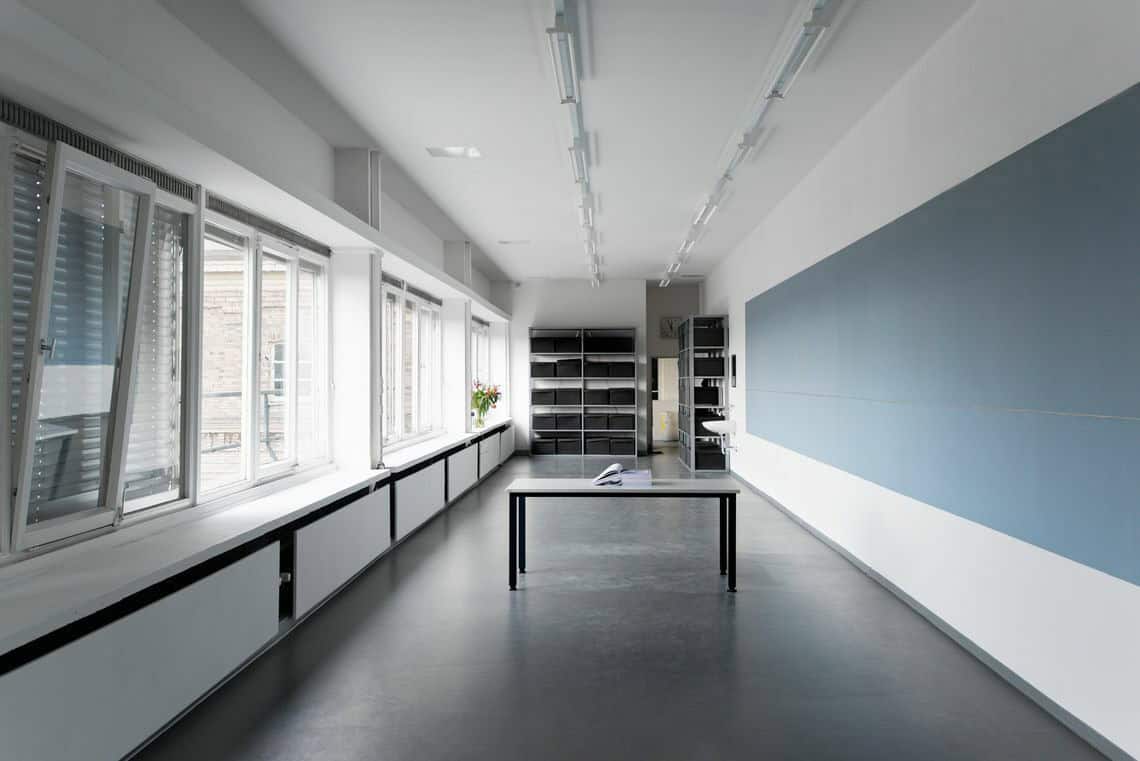 Foto: Ein länglicher, schmaler Raum mit Fensterfront. Die Wände sind weiß, der Boden schwarz. Auf der Wand ist ein Streifen aus Blauen Platten angebracht. Hinten im Raum stehen Regale, in der Mitte des Raumes steht ein Tisch, auf dem ein aufgeschlagenes Buch liegt.