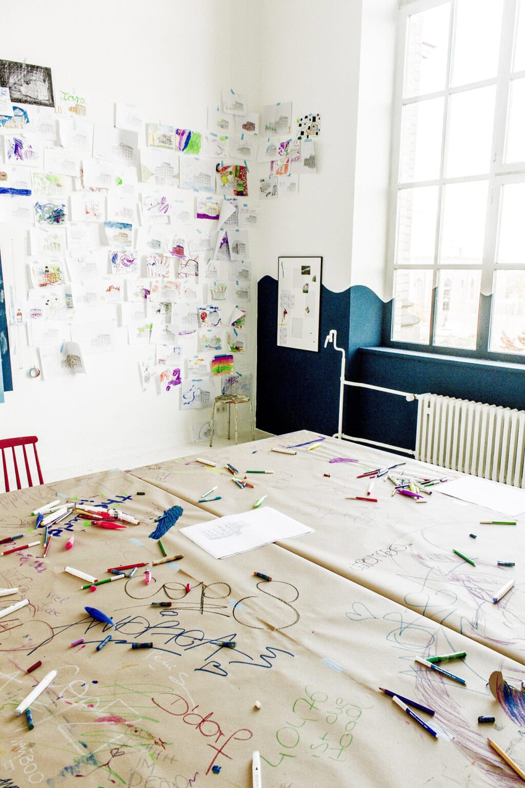 Foto: Eine große, mit Papier umwickelte Tischplatte. Auf dem Papier liegen viele Stifte und der Untergrund ist mit verschiedenfarbigen Schriftzügen bekritzelt. Dahinter eine Raumecke mit weißen Wänden und einem Fenster. Ein Teil der Wand und des Fenster ist mit dunkler, in Wellenform auslaufender Farbe bemalt. Im Raum und am Tisch stehen Stühle. An der Wand hängt eine Vielzahl von bunt bemalten Papieren.