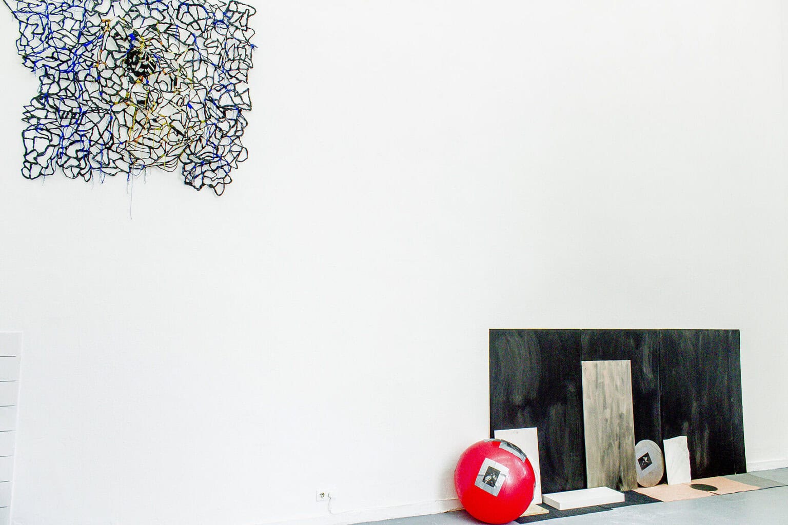 Foto: Eine weiße Wand und grauer Boden. Vor dem Wand auf dem Boden lehnt eine Gruppe von Objekten auf unterschiedlichen Materialien. An der Wand ein weiteres, netzartiges Objekt.