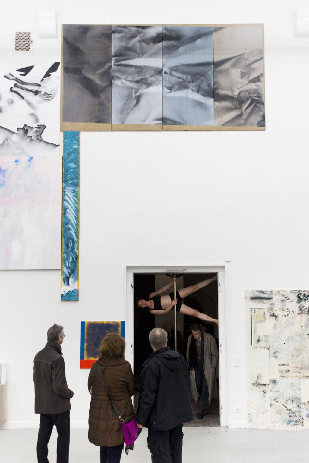 Foto: Ein Raum mit weißen Wänden, grauem Boden und einer geöffneten Tür. An den Wänden des Raumes hängen viele Flache Objekte neben- und übereinander. Im Raum stehen drei Personen. Zwischen dem Boden und der Oberkante des Türrahmens ist eine Stange montiert. Eine Person hält sich an der Stange fest und stützt sich mit den Beinen in dem Türrahmen ab, so das sie horizontal im oberen Bereich des Türrahmens schwebt. Menschen laufen in geduckter Haltung unter ihr hindurch.