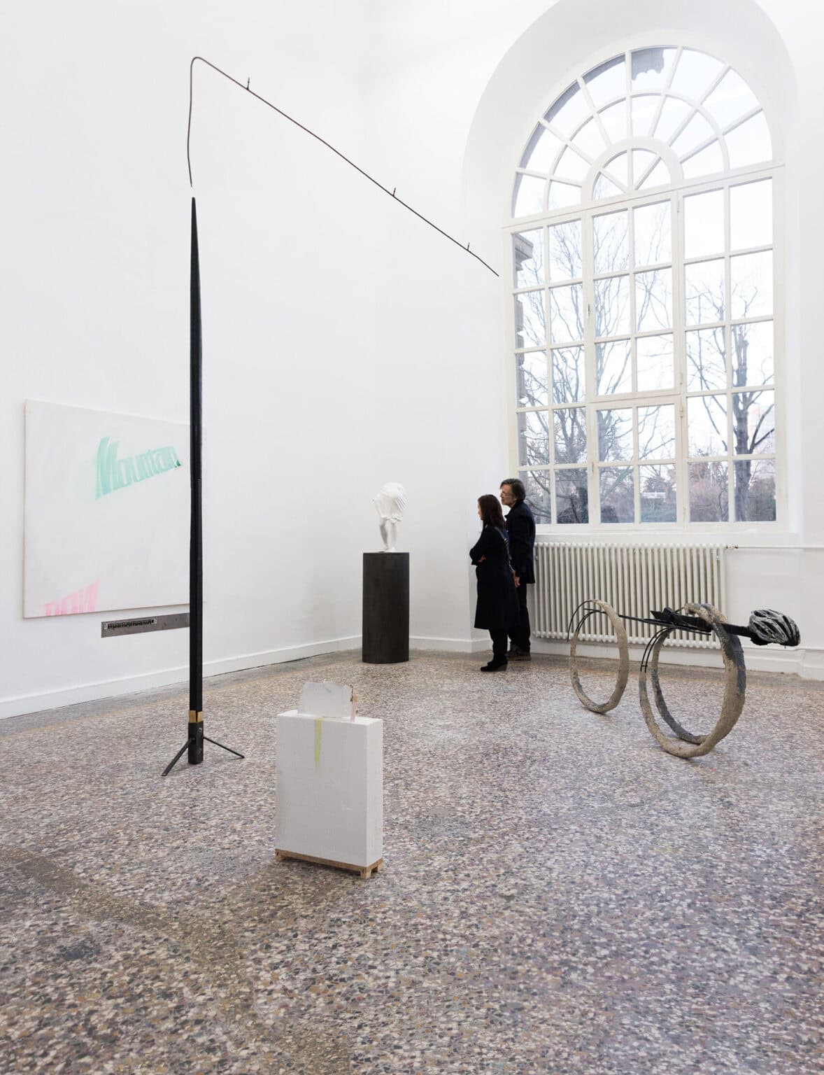 Foto: Ein Raum mit weißen Wänden, mehrfarbigem Steinboden und einem großen Fenster. An der Wand befinden sich flache Objekte. Im Raum stehen mehrere Skulpturen in unterschiedlichen Größen und Höhen. Manche von Ihnen liegen oder stehen direkt auf dem Boden, andere sind auf Sockeln installiert. Im Raum befinden sich zwei Personen.