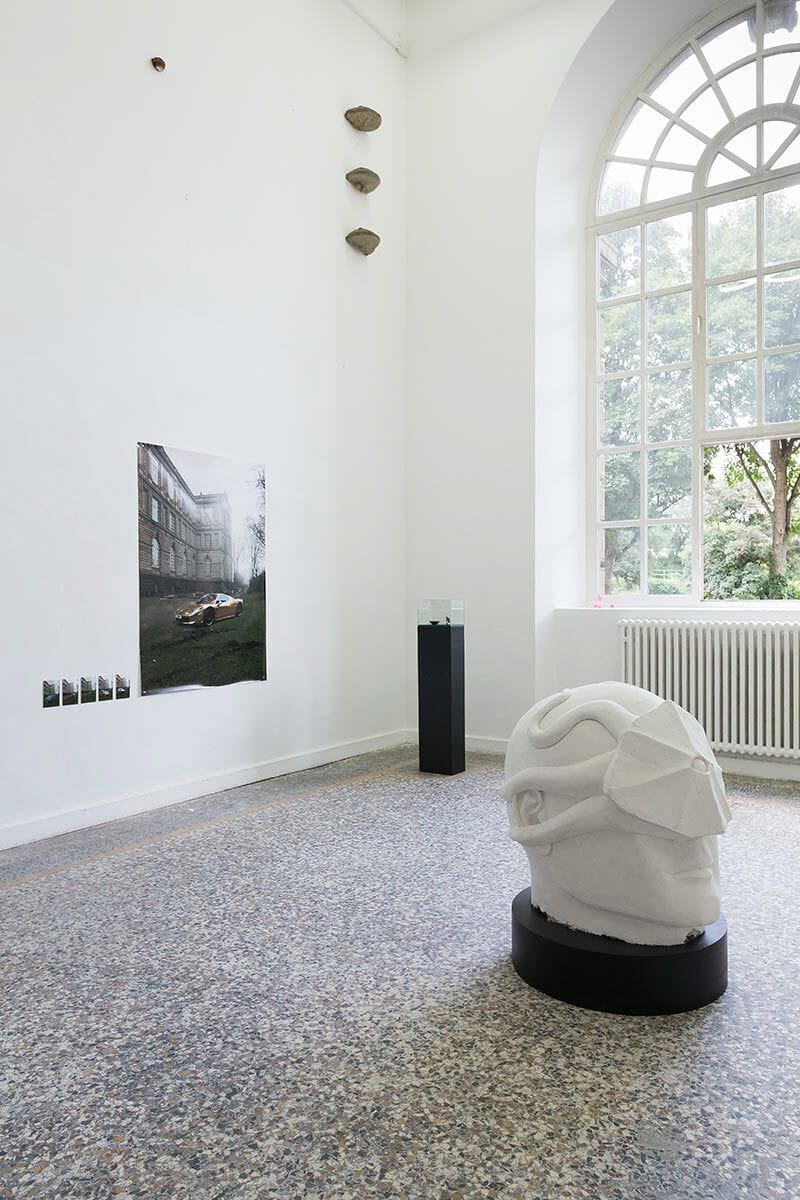 Foto: Ein Raum mit weißen Wänden und buntem Steinboden. An den Wänden hängen groß- und kleinformatige, flache Objekte auf unterschiedlicher Höhe. Im Raum stehen zwei Skulpturen.