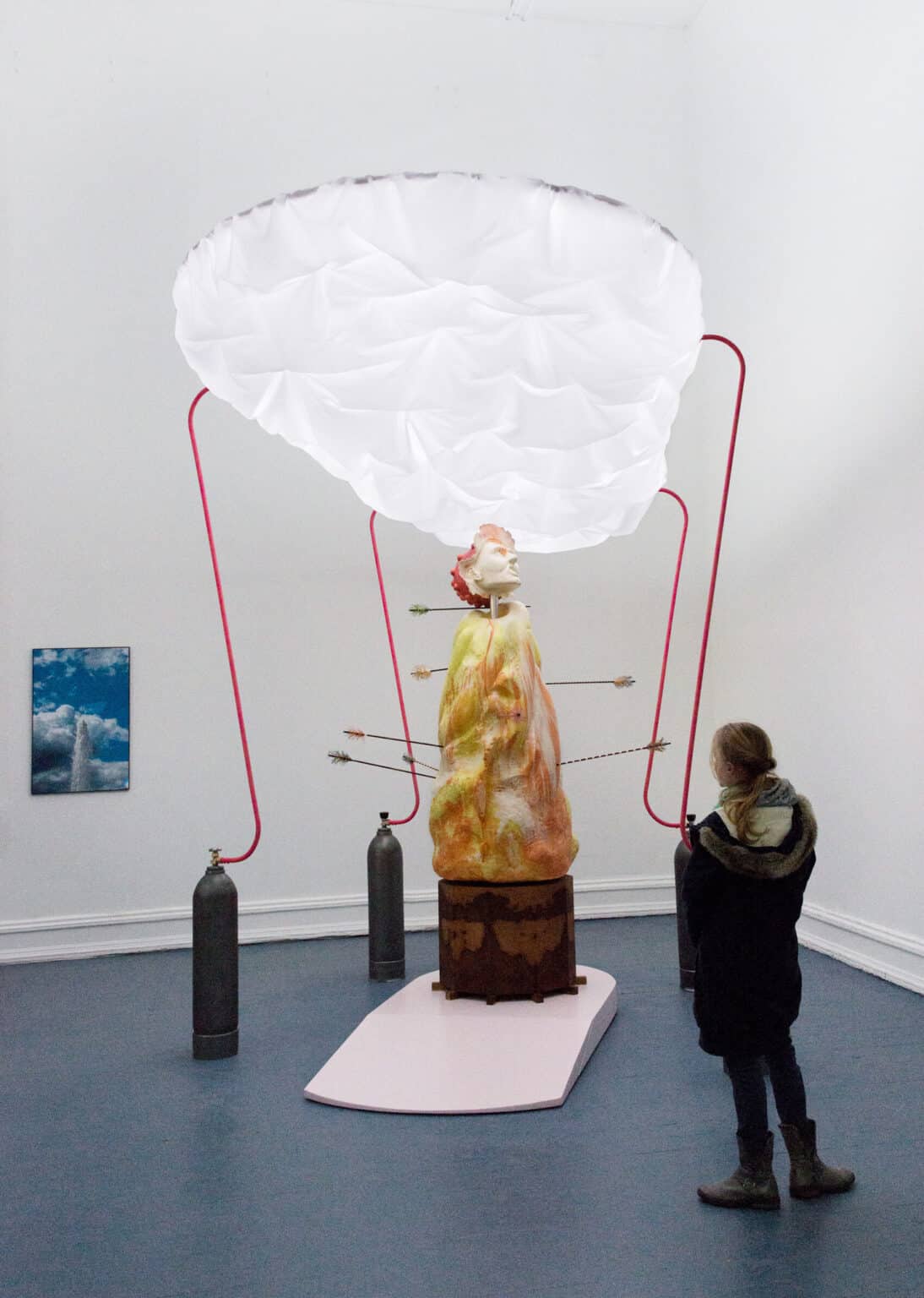 Foto: Ein Raum mit weißen Wänden und grauem Boden. Im Raum befindet sich eine große, hohe Skulptur, die aus unterschiedlichen Materialien zu bestehen scheint. An der Wand hängt ein flaches Objekt.