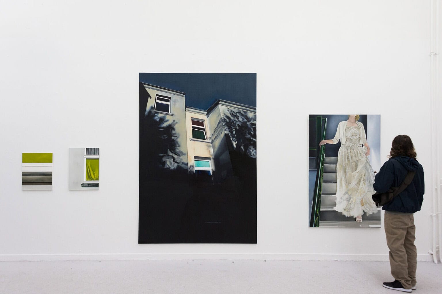 Foto: Ein Raum mit weißer Wand und hellgrauem Boden. An der Wand befinden sich Leinwandarbeiten im Klein-, Mittel- und Großformat. im Raum steht ein Mensch.