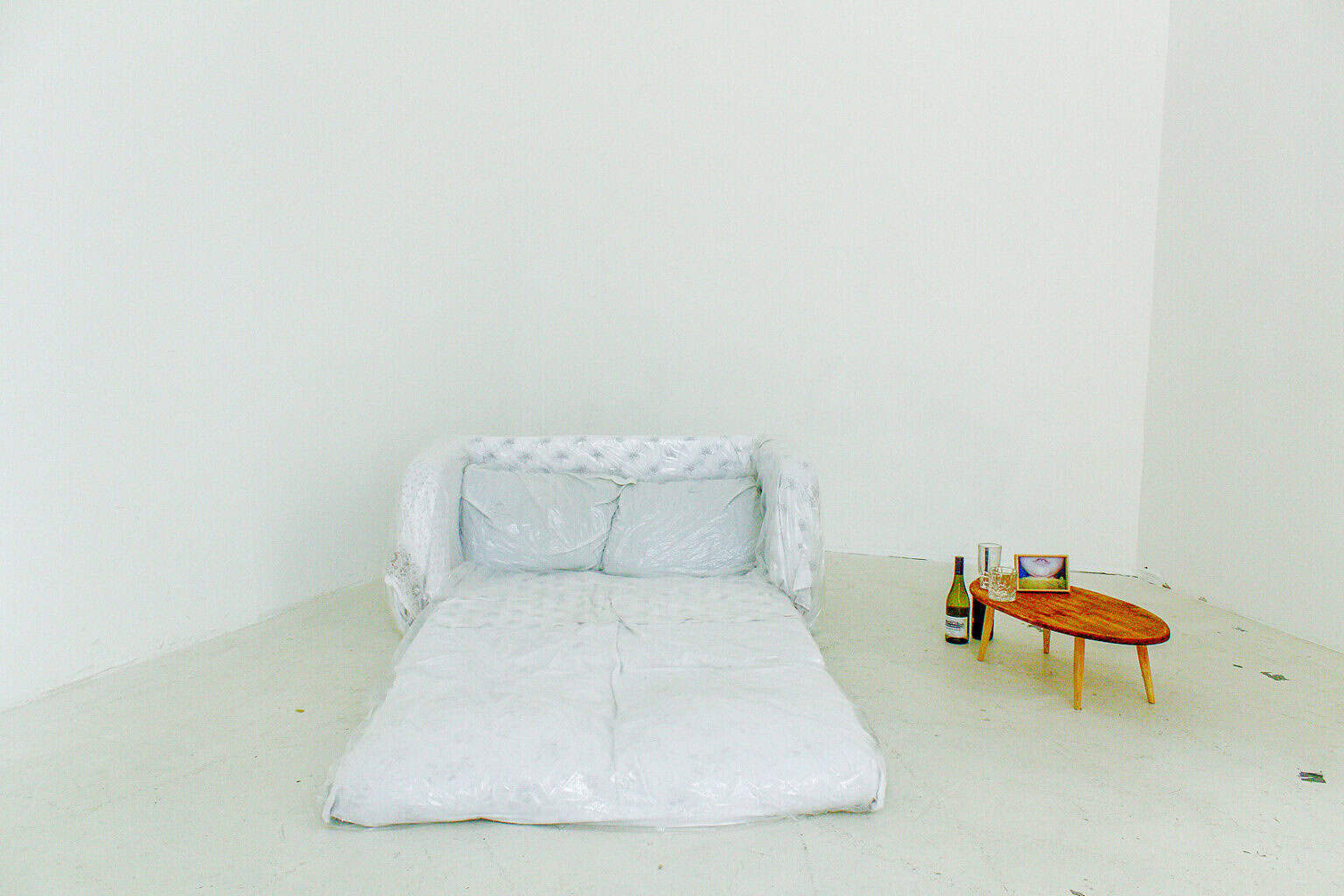 Foto: Ein Raum mit weißen Wänden und grauem Boden. Auf dem Boden des Raumes befindet sich ein gepolsterter Gegenstand, der mit einer Folie umwickelt ist. Daneben steht ein kleiner Beistelltisch. Auf und um den Tisch herum befinden sich ein Bilderrahmen, zwei Gläser und eine Flasche.