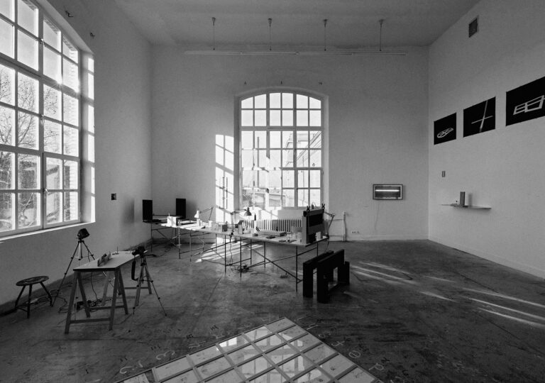 Foto: Raumansicht in schwarz weiß. Raum mit zwei großen Bogenfenstern. Im Raum und an den Wänden sind Tische und Objekte verteilt.