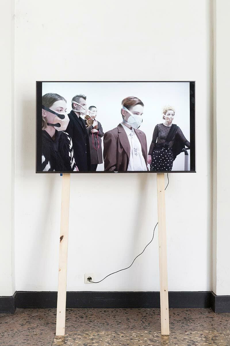 Foto: Eine weiße Wand mit schwarzer Fußleiste. Auf dem Boden stehen zwei Holzleisten, an die ein eingeschalteter Bildschirm montiert ist.