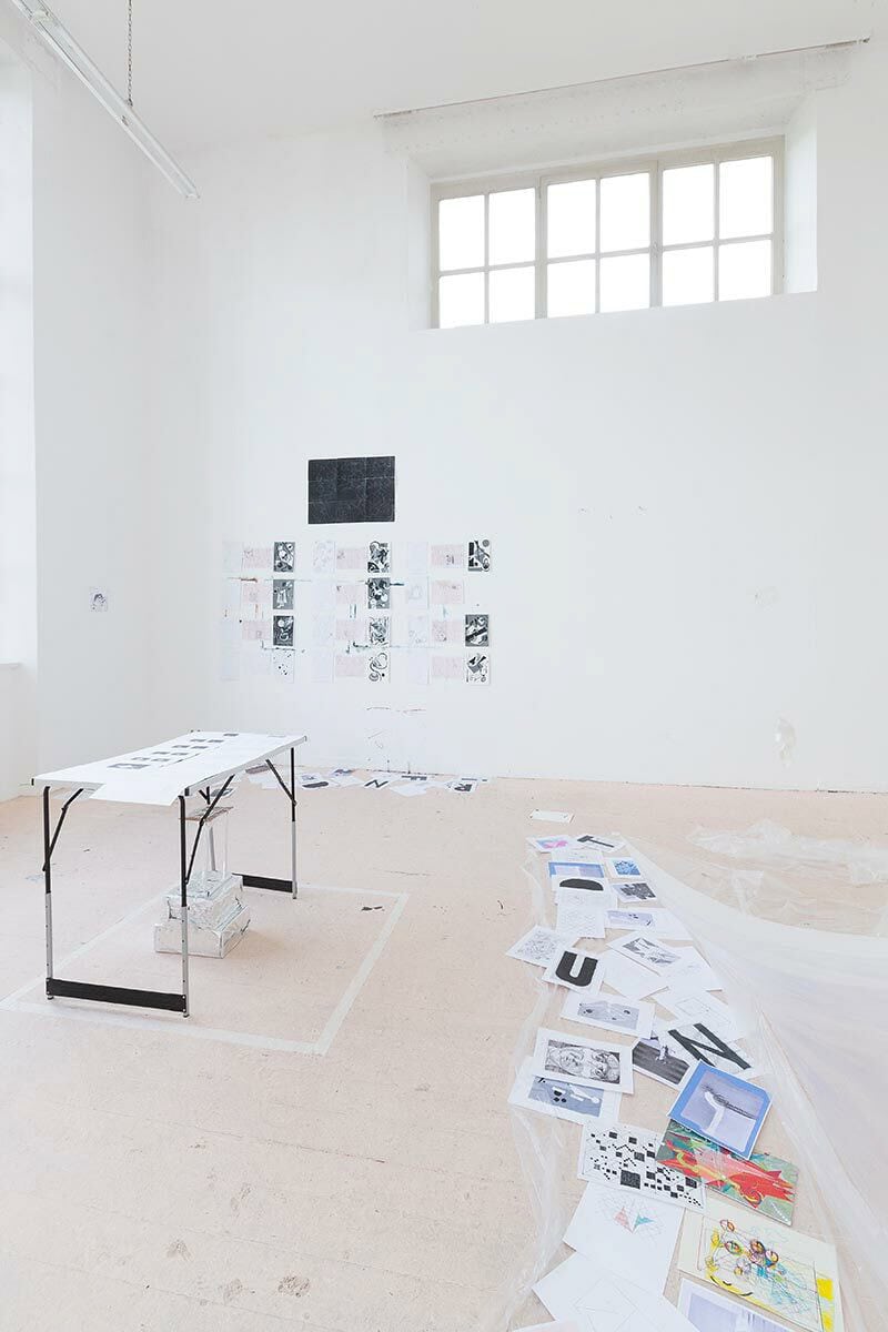 Foto: Ein Raum mit weißen Wänden, beigem Boden und kleinen Fenstern. An der Wand hängt eine Gruppe von Papieren. Auf dem Boden liegen ebenfalls Papiere und Stoff, auf dem sich Papiere befinden. Auf dem Boden ist ein Quadrat mit Tape geklebt. Innerhalb des Quadrats steht ein Tisch, auf dem Papiere liegen. Unter dem Tisch liegt ein Objekt.