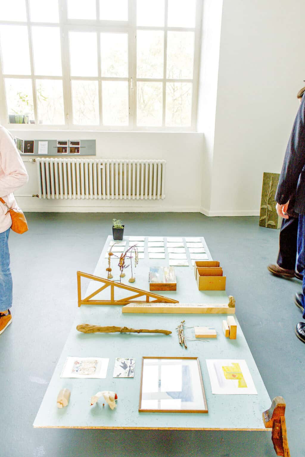 Foto: Ein Raum mit weißen Wänden, grauem Boden und einem großen Fenster. Inter der Fensterbank ist ein kleines Regal angebracht, auf dem Hefte und Objekte liegen. Auf dem Boden des Raumes steht eine große Holzplatte, auf der Zettel, gerahmte Objekte, Pflanzen und weitere Gegenstände liegen.