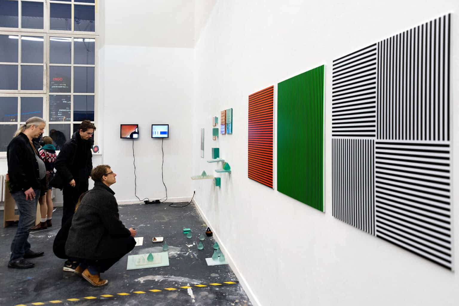 Foto: Ein Raum mit weißen Wänden, grauem Boden und einem großen Fenster. Auf dem Boden klebt eine Linie aus Absperrband. An den Wänden hängen, auf ähnlicher Nähe, zwei eingeschaltete kleine Bildschirme, Objekte und flache Objekte. Auf dem Boden liegen ebenfalls Gruppen auf kleinen Objekten. Der Boden ist mit Farbsprenklern übersäht. Im Raum befinden sich Menschen.