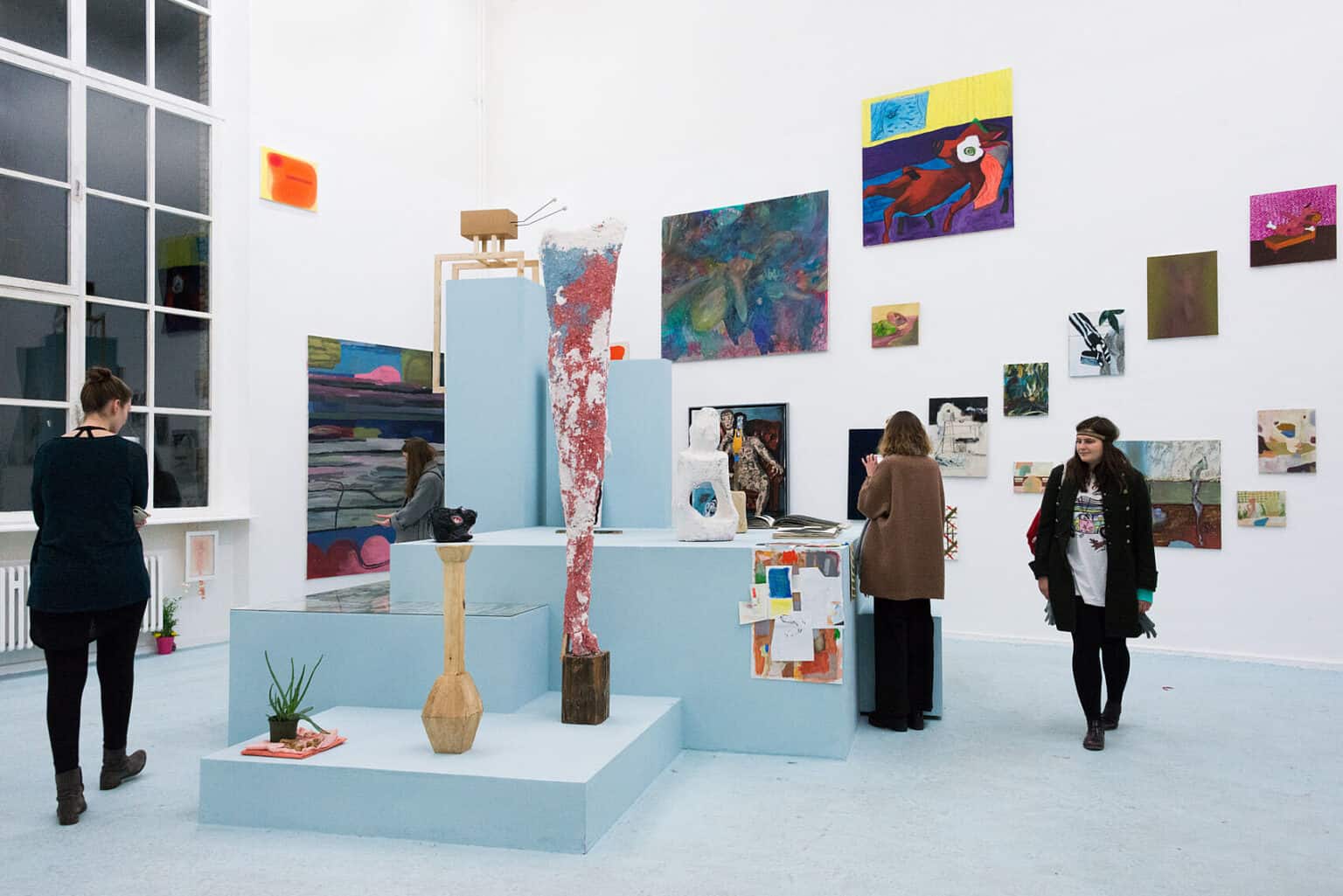 Foto: Ein Raum mit weißen Wänden und blauem Boden. An den Wänden hängen neben und übereinander viele flache Objekte in unterschiedlichen Formaten auf unterschiedlichen Höhen. Im Raum ist ein verschachteltes, ebenfalls hellblaues Podest installiert, auf dem Objekte und Hefte stehen und liegen. Durch den Raum bewegen sich Menschen.