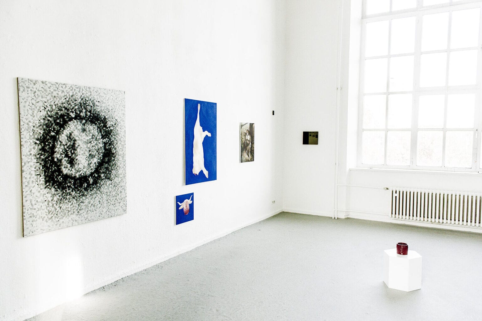 Foto: Ein Raum mit weißen Wänden, grauem Boden und großem Fenster. Im Raum befindet sich ein niedriger, sechseckiger, weißer Sockel, auf dem sich ein Objekt befindet. An der Wand hängen, auf unterschiedlicher Höhe, Leinwandarbeiten im Klein- und Mittelformat.