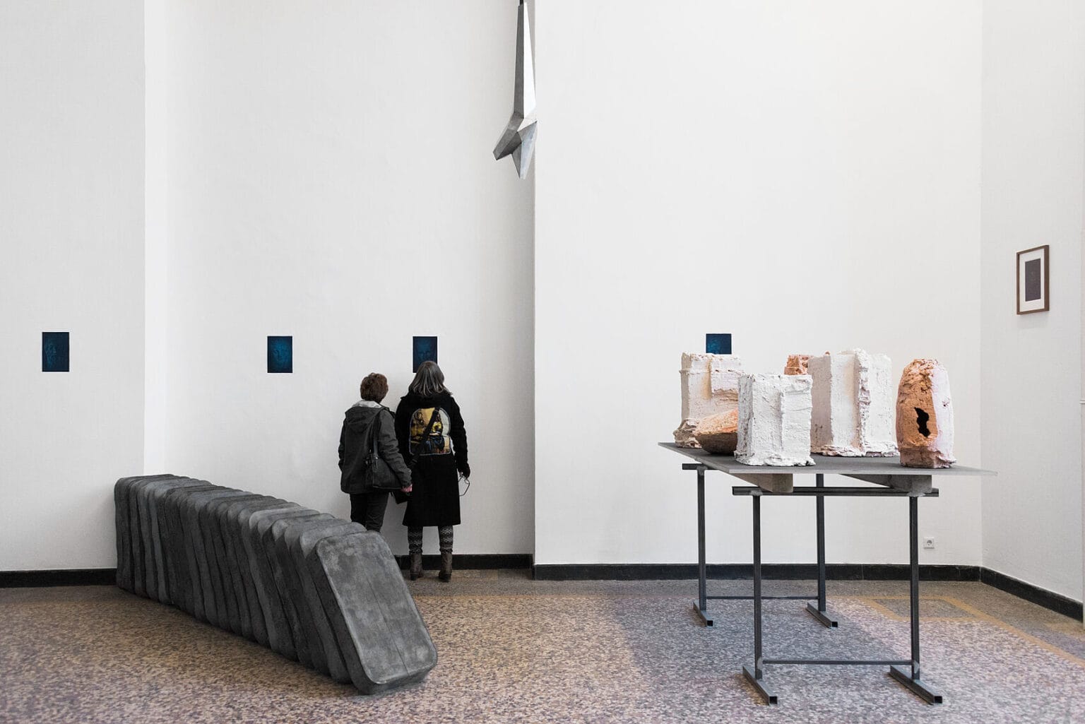 Foto: Ein Raum mit weißen Wänden und mehrfarbigem Steinbonde. An den Wänden gerahmte und ungerahmte Objekte im Kleinformat. Auf dem Boden eine Reihe rechteckiger Objekte, die aneinander Lehnen. Daneben eine große Skulptur. Von der Decke hängt ein Objekt. Im Raum stehen zwei Menschen.